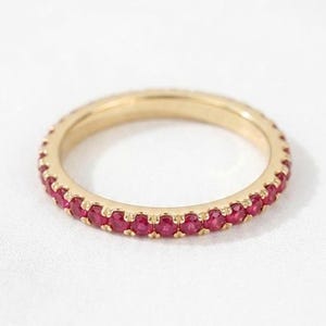 Könnte beinhalten: Ein Goldring mit einer durchgehenden Reihe kleiner, runder, leuchtend roter Rubine, die in eine Fassung aus poliertem Gelbgold eingefasst sind. Der Ring hat ein schmales Profil, wobei die Edelsteine den gesamten Umfang umschließen.