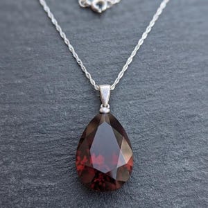 Könnte beinhalten: Ein tropfenförmiger, dunkelroter Edelsteinanhänger an einer dünnen, silberfarbenen gedrehten Kette. Der facettierte Stein hat einen tiefen Burgunderfarbton mit geometrischen Schliffen, die das Licht reflektieren, gehalten von einer silbernen Öse.