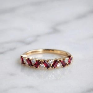 Könnte beinhalten: Ein Goldring mit einem abwechselnden Muster aus tiefroten Rubinen im Baguetteschliff und klaren, runden Diamanten im Brillantschliff. Die Steine sind in einer durchgehenden Reihe auf der oberen Hälfte des polierten Goldrings angeordnet.