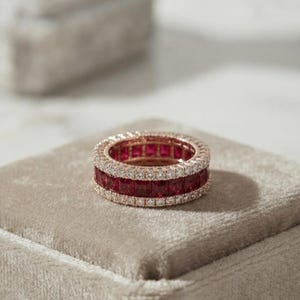 May include: A rose gold eternity band featuring a central row of square-cut deep red rubies flanked by two outer rows of small, round, clear pavé-set diamonds. The ring has a continuous, uniform design with a polished metal finish throughout.