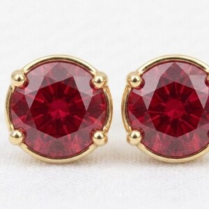 Pode incluir: Um par de brincos redondos com pedras vermelhas facetadas em engastes de metal dourado polido. Cada pedra circular é fixada por quatro pequenas garras, criando um design clássico que destaca a cor carmesim profunda das pedras.