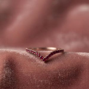 May include: A V-shaped ring crafted from rose-toned metal, featuring a series of small, round, deep red gemstones set along the entire chevron-style band. The ring displays a polished finish and a delicate, symmetrical design.