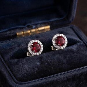 May include: A pair of round stud earrings featuring a deep red center stone surrounded by a halo of small clear stones. The earrings are set in a metallic frame and are positioned inside a dark blue velvet-lined jewelry box with a hinged lid.