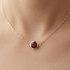 Pode incluir: Um colar de corrente dourada delicada com uma única pedra preciosa redonda e facetada de cor vermelho profundo, engastada num aro de ouro liso e polido. O colar está centrado com a pedra como ponto focal, ligada a elos dourados finos.