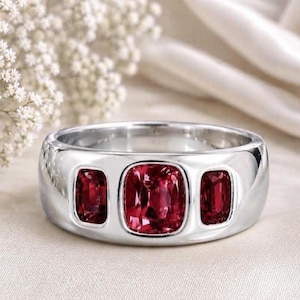 May include: A polished silver-toned band ring featuring three bezel-set, cushion-cut deep red gemstones. The center stone is larger than the two flanking stones, all set horizontally within the smooth, reflective metal surface of the ring.
