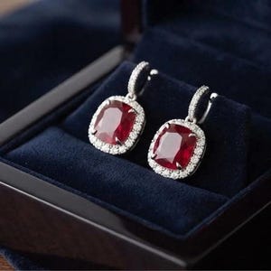 May include: A pair of drop earrings featuring large, cushion-cut deep red gemstones surrounded by a halo of small, clear white stones. The earrings are set in a metallic frame with a pavé-set bail, presented inside a dark blue velvet-lined box.
