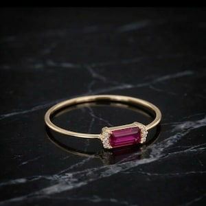 May include: A thin gold ring featuring a rectangular baguette-cut red gemstone set horizontally. The centre stone is flanked on both sides by two small, clear round accent stones, creating a delicate and refined piece of jewellery.