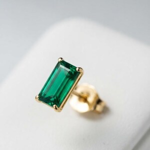 May include: A single stud earring featuring a rectangular, emerald-cut green gemstone set in a gold-toned metal prong setting. The earring includes a matching gold-toned metal post and a secure butterfly-style backing mechanism.
