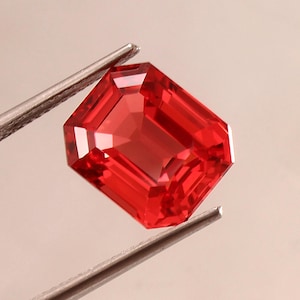 May include: A vibrant, rectangular, emerald-cut gemstone with a deep red hue. The stone features multiple facets that reflect light, creating a brilliant appearance. It is held by a pair of silver-toned metal tweezers, showcasing its clarity and color.