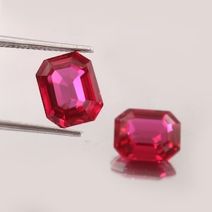 Peut inclure: Deux pierres précieuses rouge vif de taille émeraude, dotées de multiples facettes qui reflètent la lumière. L'une des pierres est tenue par une pince en métal argenté, tandis que l'autre repose à proximité, créant un reflet net en dessous.