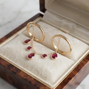 May include: Two gold rings connected by a delicate gold chain featuring five dangling red gemstones. Three of the stones are pear-shaped and two are round, all set in gold bezels. The jewellery is displayed inside a polished wooden box with cream lining.