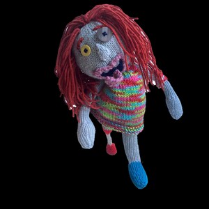 Puede incluir: Muñeca zombi tejida a mano con cabello de hilo rojo, un ojo de botón y un suéter a rayas. La muñeca tiene un pie azul y un trozo rojo colgante. Sobre un fondo negro.