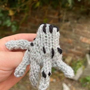 Peut inclure: Petit jouet en forme de poulpe, tricoté à la main, gris et noir. Le poulpe tricoté a des tentacules déployées et une surface texturée. Le jouet est fait de fil et est tenu dans une main.