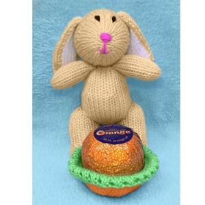 Puede incluir: Un conejito de punto beige sosteniendo una naranja de chocolate en una cesta de crochet verde. La naranja de chocolate está envuelta en papel de aluminio naranja y azul con el texto "Chocolate Orange" y "Real Orange Oil".