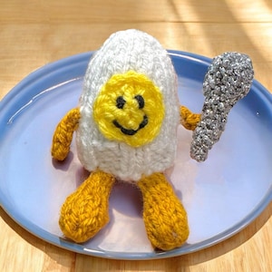 May include: A whimsical, handmade knitted egg character with a smiling yellow face, yellow arms and feet, and a silver spoon. The egg is white with a yellow yolk and black smile. The character is on a light blue plate.