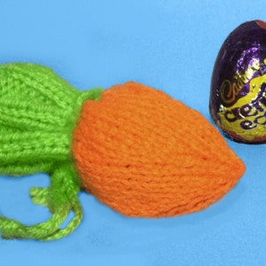 Peut inclure: Une carotte orange tricotée avec un dessus vert et une ficelle verte. La carotte est posée sur une surface bleue à côté d'un œuf en chocolat enveloppé de papier d'aluminium violet et or.