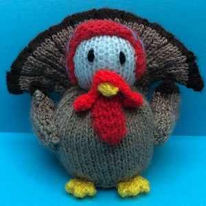 May include: A knitted turkey toy with a brown and gray body, red wattle, and yellow feet. The turkey has a white head with black eyes and a blue and white knitted hat.