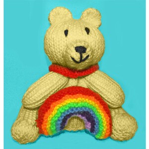 Puede incluir: Un oso de peluche beige tejido sosteniendo un arcoíris tejido de colores arcoíris en sus patas. El oso tiene una bufanda roja alrededor del cuello.