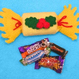 Puede incluir: Un cracker navideño de punto amarillo con una cinta roja y una decoración de acebo verde. El cracker está lleno de tres barras de chocolate: Bounty, Mars y Crunchie.