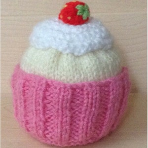 May include: A knitted cupcake toy with pink, white, and light yellow frosting. The cupcake has a red and green felt strawberry on top.
