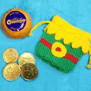 May include: A small green and yellow knitted bag with a red stripe and a yellow circle in the center. The bag is sitting on a blue surface next to a chocolate orange and four gold coins.