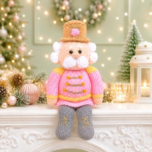 Pode incluir: Um boneco quebra-nozes de tricô com um chapéu bege com um detalhe rosa, uma jaqueta rosa claro com acabamento amarelo e botões dourados, cabelo e barba brancos, e botas cinzas com detalhes em ponto cruz dourado na frente de cada pé.