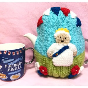 May include: A knitted teapot cozy featuring a cartoon figure of the Queen in a white dress and a blue sash. The cozy is decorated with red, white, and blue bunting and red flowers. The text "Celebrating The Queen's Platinum Jubilee" is printed on a blue mug.