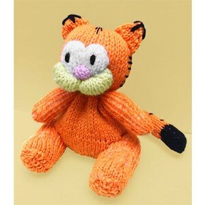 Puede incluir: Juguete de peluche de punto naranja con forma de gato con ojos negros, nariz rosa y boca verde. El juguete está sentado sobre una superficie amarilla.