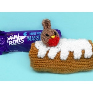 Puede incluir: Un robin tejido posado sobre un pastelito de rollo mini tejido en marrón y blanco. El pastel está al lado de un envoltorio morado y blanco para un bocadillo de chocolate Cadbury Mini Rolls.