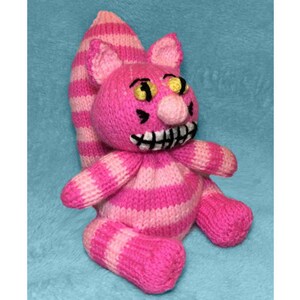 May include: A knitted Cheshire Cat toy in pink and white stripes. The cat has a wide grin with black teeth, yellow eyes, and a long striped tail. The plush toy is set against a blue background.