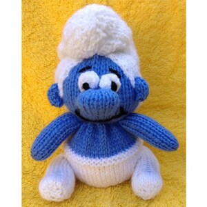 Könnte beinhalten: Ein blaues, gestricktes Plüschtier mit einer weißen, gestrickten Mütze und Windel. Das Spielzeug hat ein lächelndes Gesicht mit schwarzen Augen und einem schwarzen Mund.