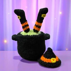 May include: A black crocheted cauldron with green liquid, with two black and orange striped witch legs protruding. A small black witch hat with an orange band and yellow buckle is next to the cauldron. Halloween decoration.