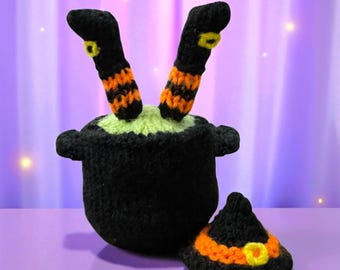 Op de afbeelding: Een zwart gehaakte ketel met groene vloeistof, met twee zwart-oranje gestreepte heksenbenen die uitsteken. Een kleine zwarte heksenhoed met een oranje band en gele gesp staat naast de ketel. Halloween-decoratie.