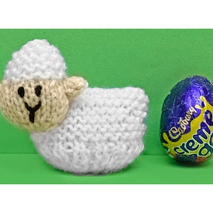 Op de afbeelding: Een wit gebreid lammetje dat een paars en geel Cadbury Creme Egg vasthoudt.