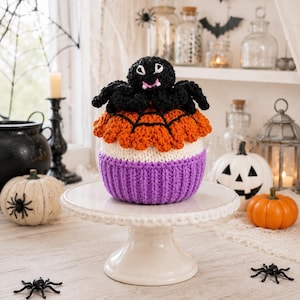 May include: A crocheted cupcake-shaped decoration featuring a black spider with white eyes and pink fangs on top. The cupcake has an orange top layer with a black web pattern, a white middle band, and a textured purple base, sitting on a white stand.