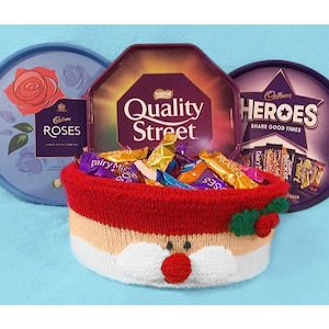 May include: A knitted Santa Claus basket filled with assorted chocolate sweets. The basket is red and white with a green holly sprig. The sweets are in boxes labelled "Roses", "Quality Street", and "Heroes".