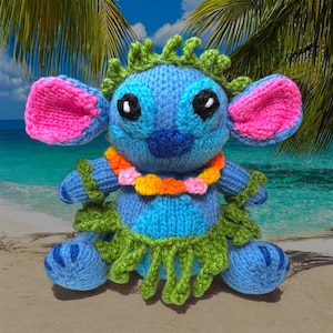 May include: A blue knitted Stitch plush toy with pink ears, black eyes, and a blue nose. It wears a green grass skirt and a flower lei. The background shows a beach scene with palm trees and blue water.