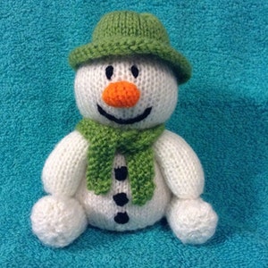 May include: A white knitted snowman wearing a green hat and scarf. The snowman has black eyes, a black mouth, and an orange carrot nose.