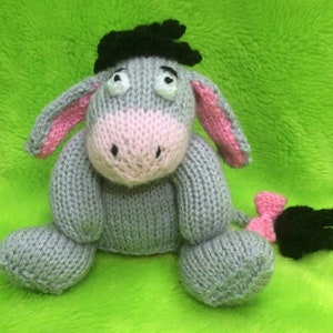 Puede incluir: Un juguete de peluche de burro gris tejido con pelo negro, orejas rosas y cola rosa. El juguete tiene una expresión dulce con ojos grandes y una nariz pequeña.