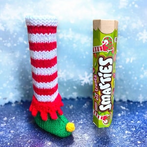 May include: A red and white striped knitted elf boot with a green foot and a yellow toe. The boot is next to a box of Smarties elf treats.