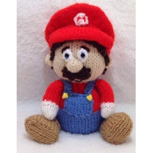 May include: A knitted plush toy of the video game character Mario. He is wearing a red hat with a white 'M' on it, a red shirt, blue overalls, and brown shoes.