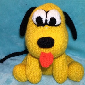 Könnte beinhalten: Ein gelber, gestrickter Plüschhund mit schwarzen Ohren und einem schwarzen Schwanz. Der Hund hat große weiße Augen mit schwarzen Pupillen, eine schwarze Nase und eine rote Zunge.