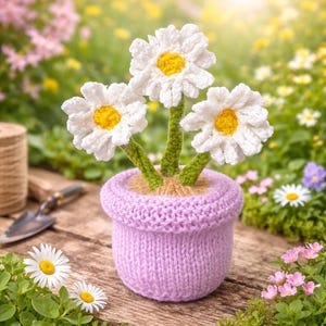 Può includere: Una composizione floreale di margherite lavorate a maglia in un vaso lilla. Le margherite hanno petali bianchi e centri gialli, con steli verdi. Il vaso è di colore viola chiaro e poggia su una superficie di legno. Lo sfondo è una scena di giardino sfocata.