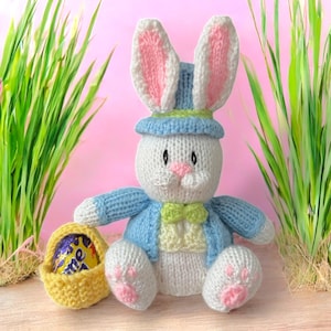 May include: A white knitted Easter bunny wearing a blue hat with a green trim and a green bow tie. The bunny is holding a yellow knitted basket with a chocolate egg inside.