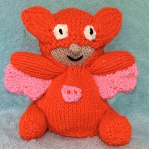 May include: A knitted plush toy in the shape of a bat wearing a red and pink costume. The bat has large eyes and a small pink nose.