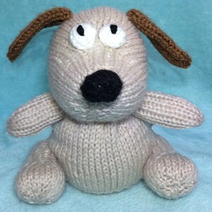 May include: A knitted beige dog toy with brown ears and black eyes. The dog has a black nose and is sitting on a blue surface.