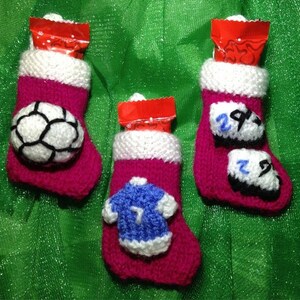 May include: Three pink knitted Christmas stockings with white trim. Each stocking has a different sports-themed decoration: a soccer ball, a jersey with the number 7, and two soccer cleats with the number 29.