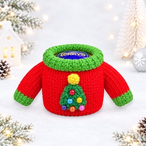 May include: A red crocheted jumper cover for a Terry's chocolate orange, featuring a green ribbed collar and cuffs. The front displays a green crocheted Christmas tree with a yellow pom-pom topper and small, colourful round bead ornaments.