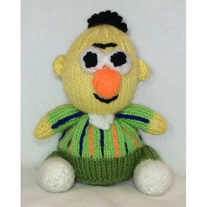 Op de afbeelding: Een gebreid pluche speelgoed met een geel lichaam, een groen en oranje gestreept shirt, zwart haar en een grote oranje neus. Het speelgoed heeft witte ogen en een zwarte mond.