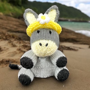Pode incluir: Um brinquedo de burro de malha cinzenta com cascos pretos, usando uma faixa de malha amarela com uma margarida branca no topo. O burro está sentado numa praia de areia.